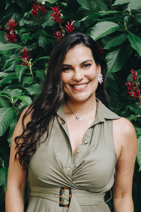 Remedial Massage Therapist, Catalina Corrales, smiling and standing in front of a lush green plant with red flowers.